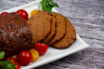Vegan meat alternative- seitan seasoned with spiced, glazed with BBQ sauce. White plate, sliced, wooden table.