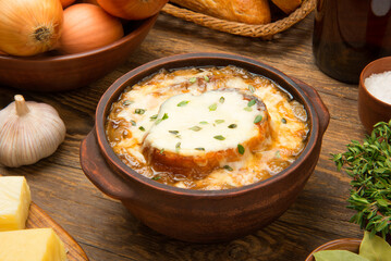 French onion soup in a brown ceramic bowl on a rustic wooden table with ingredients.  - 676101321