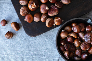 Brown roasted chestnuts on black wooden board and in pan, on gray linen tablecloth, traditional winter, Christmas holiday treat