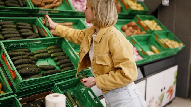 Blonde woman shopping for fresh cucumbers in supermarket, selecting produce, concept of healthy eating and grocery shopping