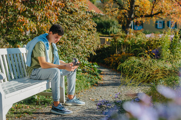 Fototapeta premium A Teenager Sits On A Bench In The Autumn Park Drinks Coffee From A Thermo Mug And Looks Into A Phone. Portrait Of Handsome Cheerful Guy Sitting On Bench Fresh Air Using Device Browsing Media Smm