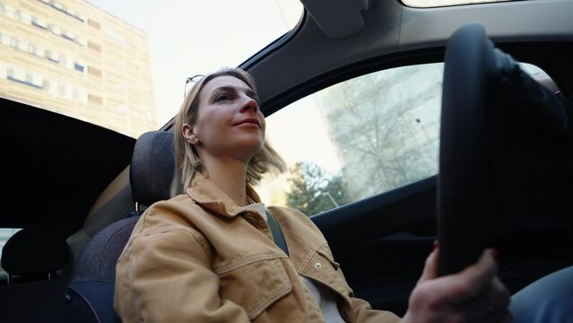 Blonde woman attentively driving car, looking ahead, skyscrapers and clear sky in view, reflecting focused urban driving and navigation
