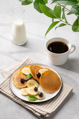 Delicious banana pancakes with fresh fruit slices, berries, mint and honey on a light background with a cup of black coffee, milk and green branch.