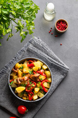 Fried pork meat with potatoes, onions and peppers. National homemade Georgian dish ojakhuri in a bowl on a dark background with spices, fresh tomato and herb.