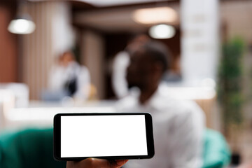 Selective focus of white screen phone, people relaxing in lounge area at hotel lobby. Person holding smartphone with blank template on display with isolated copyspace, modern resort. Close up.