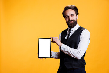 Young waiter holding tablet with white screen on camera, presenting isolated empty template against yellow background. Professional employee shows device with blank mockup display.