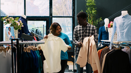 African american couple looking at trendy clothes, shopping for casual wear in modern boutique. Cheerful customers checking outfit material, buying stylish merchandise for new wardrobe. Fashion