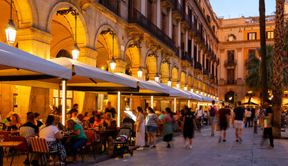 Citizens and tourists stroll and relax in cafe in light of streetlight on Royal square in Barcelona. Atmosphere of summer evening in Gothic quarter - obrazy, fototapety, plakaty