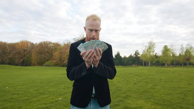 Bright image of a red-haired man with beard holding a bunch of dollars, looking extremely surprised. Vast territory with green lawn and autumn trees. High quality 4k footage