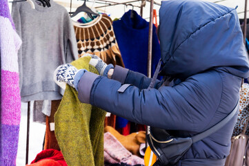 Woman choosing clothing in a second hand store. Various vintage sweaters hang on clothing rack. Thrifting and sustainability in clothing concept