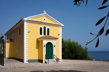 Barbati, Corfu - August 20, 2022: Old small yellow church or Chapel with a green door on the shore of a calm deep sea on Corfu, Greece.