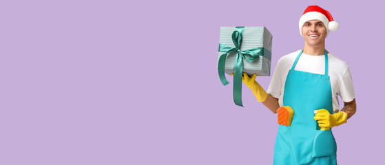 Male cleaner with Christmas gift on lilac background with space for text