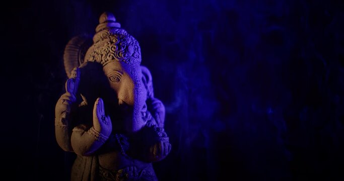Ganesha idol made of sandstone on a black background at night. There are gradually alternating colorful light colors. There is smoke from the incense that people burn to worship.