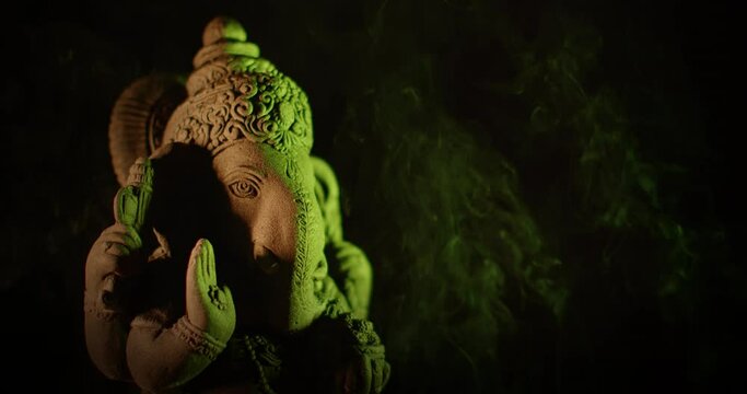 Ganesha idol made of sandstone on a black background at night. There are gradually alternating colorful light colors. There is smoke from the incense that people burn to worship.