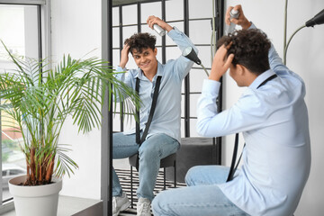 Handsome young man applying hair spray on his curly hair near mirror - obrazy, fototapety, plakaty
