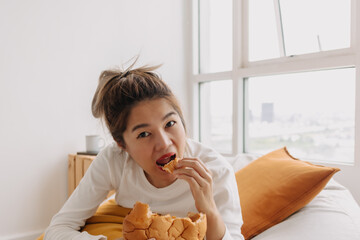 Asian woman eating bread and coffee breakfast sits on bean bag in living.