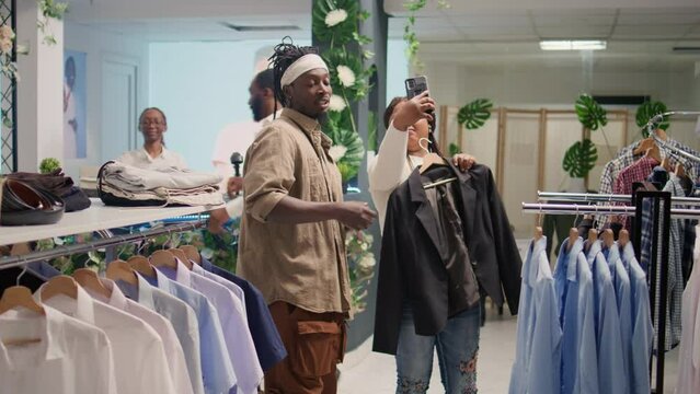 African american man and his girlfriend looking for cheap clothes in second hand clothing shop, unhappy with ugly blazer. Woman with her boyfriend in discount store looking to take pics