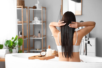 Beautiful young woman applying coconut oil onto her hair in bathroom, back view - obrazy, fototapety, plakaty