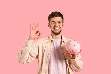 Young bearded man with piggy bank showing OK on pink background