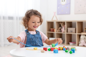 Motor skills development. Little girl playing with wooden pieces and string for threading activity at table indoors