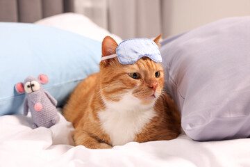 Cute ginger cat with sleep mask and crocheted mouse resting on bed