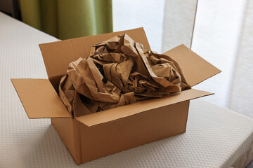 Open cardboard box with crumpled brown paper filler on white mattress indoors