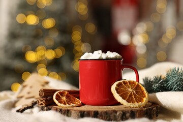 Cup of cocoa with marshmallows, cinnamon sticks, dry orange slices and Christmas decor on table against festive lights, closeup
