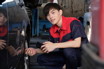 Young male Asian professional automotive mechanic worker checks tire pressure by smartphone application at a car garage, expert in maintenance vehicle service, and fixing occupations auto industry.