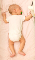 A baby in a white bodysuit sleeps with a pacifier in the crib.