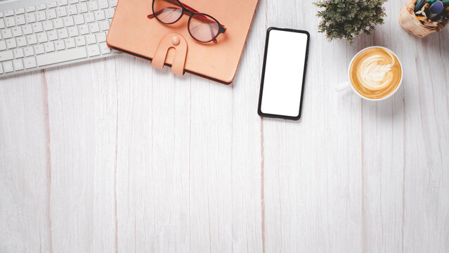 White wooden desk with blank screen smart phone, keyboard, eyeglass, notebook and cup of coffee, Top view flat lay with copy space, Blank screen for product display.