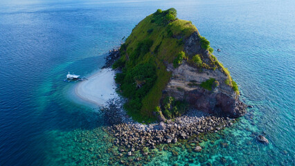 Tropical island in the open sea. Aerial view of a small rocky uninhabited island with a small sandy beach among a coral reef. A small boat docked on the island's beach. Sombrero Island, Anilao, Philip