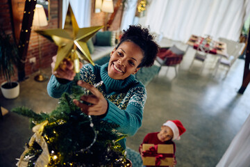 Happy black mother putting star on top of Christmas tree while decorating home with her daughter.