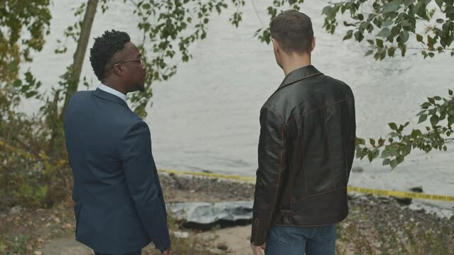 Medium back view shot of two multiethnic crime investigators having discussion while looking at murder scene and corpse in black plastic bag at river bank behind yellow barricade tape