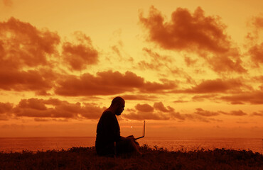 Side view of man silhouette using and looking at laptop computer during sunset over the ocean. male...