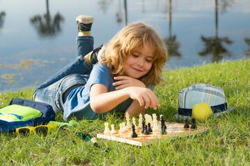 Kid playing chess game in backyard, laying on grass. Concentrated child play chess. Kid playing...