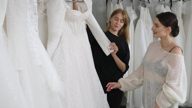 A saleswoman advises a young bride in a wedding shop. The consultant girl shows the wedding dress to the future bride. Wedding dresses are hanging on hangers.