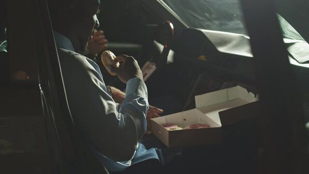 Medium shot of two ethnically diverse policemen sitting in dark car at night on mission eating donuts and looking through suspect profile sheet