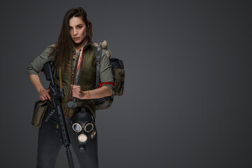 Middle Eastern-looking woman dressed in survivalist clothing posing with a rifle against a gray...