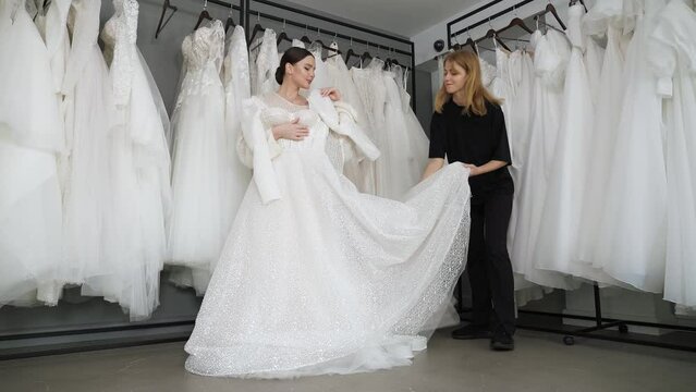 A beautiful bride chooses a wedding dress in a wedding salon, a consultant helps her. Wedding dresses are hanging on hangers. Lots of dresses. Preparation for the wedding. Wide shot.