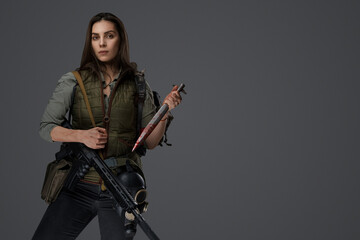 Naklejka premium Middle Eastern-looking woman dressed in survivalist clothing posing with a rifle against a gray background, portraying strength and resilience