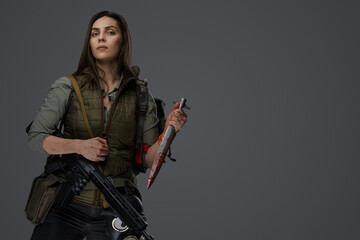 Middle Eastern-looking woman dressed in survivalist clothing posing with a rifle against a gray background, portraying strength and resilience