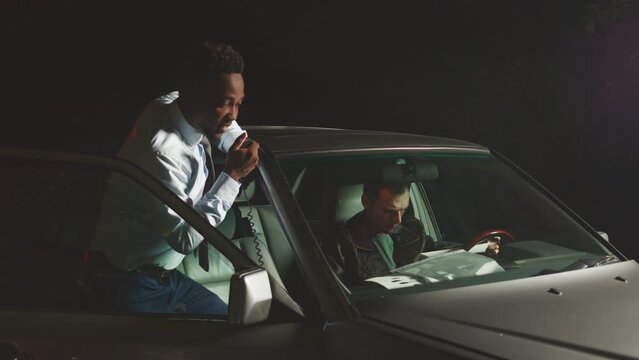 Medium shot of African American male police officer speaking in radio transmitter while standing near car at night trying to persecute criminal with colleague sitting at driver seat