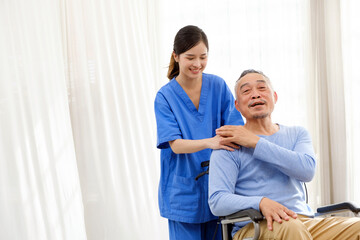 The caregiver therapist stands with an Asian senior sitting in a wheelchair and touches their hands...