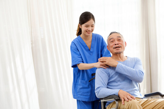 The caregiver therapist stands with an Asian senior sitting in a wheelchair and touches their hands on senior shoulder together. The nursing home facilitates a support group. Image with copy space