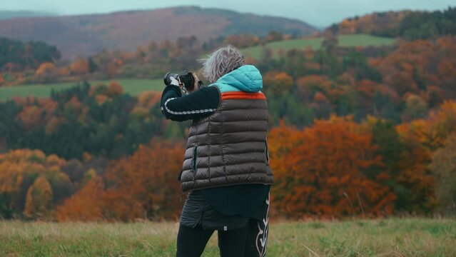 Medium shot of a female Photographer taking pictures of beautiful autumn natures with bright orange colors with her camera  and walking through the field during a windy day in slow motion.Sony FX3, 4K