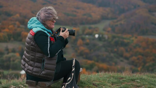 Female Photographer sitting on a bench taking pictures with her camera and looking around while wind is blowing up her grey hair, surrounded by beautiful autumn nature with colourful orange trees.