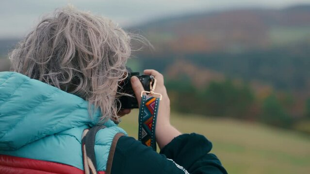 Close up of grey haired woman Photographer from behind taking pictures of colourful orange autumn natures with her camera and looking around during a cold windy day in slow motion. Sony FX3 in 4K