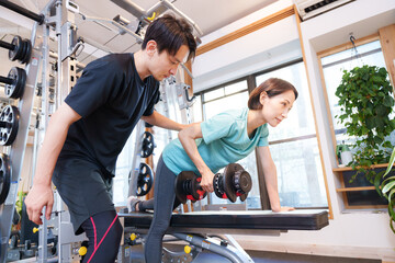 スポーツジムで運動する女性とトレーナー（ダンベル）