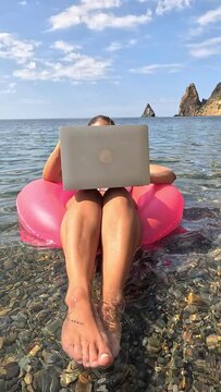 Woman freelancer works on laptop swimming in sea on pink inflatable ring. Happy tourist in sunglasses floating on inflatable donut and working on laptop computer in calm ocean. Remote working anywhere
