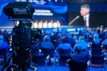 Fotobehang Video camera capturing global business conference in illuminated blue auditorium at export forum © Anton Gvozdikov
